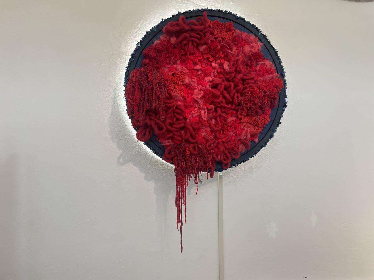 Foto dell'opera di Lisa Martignoni dove degli intrecci di lana rossa escono da un cerchio a mosaico blu.